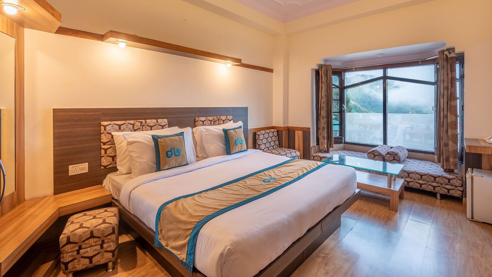 A hotel room with a large bed, a window overlooking a mountain view, and a seating area - DLS Meghavan Resort